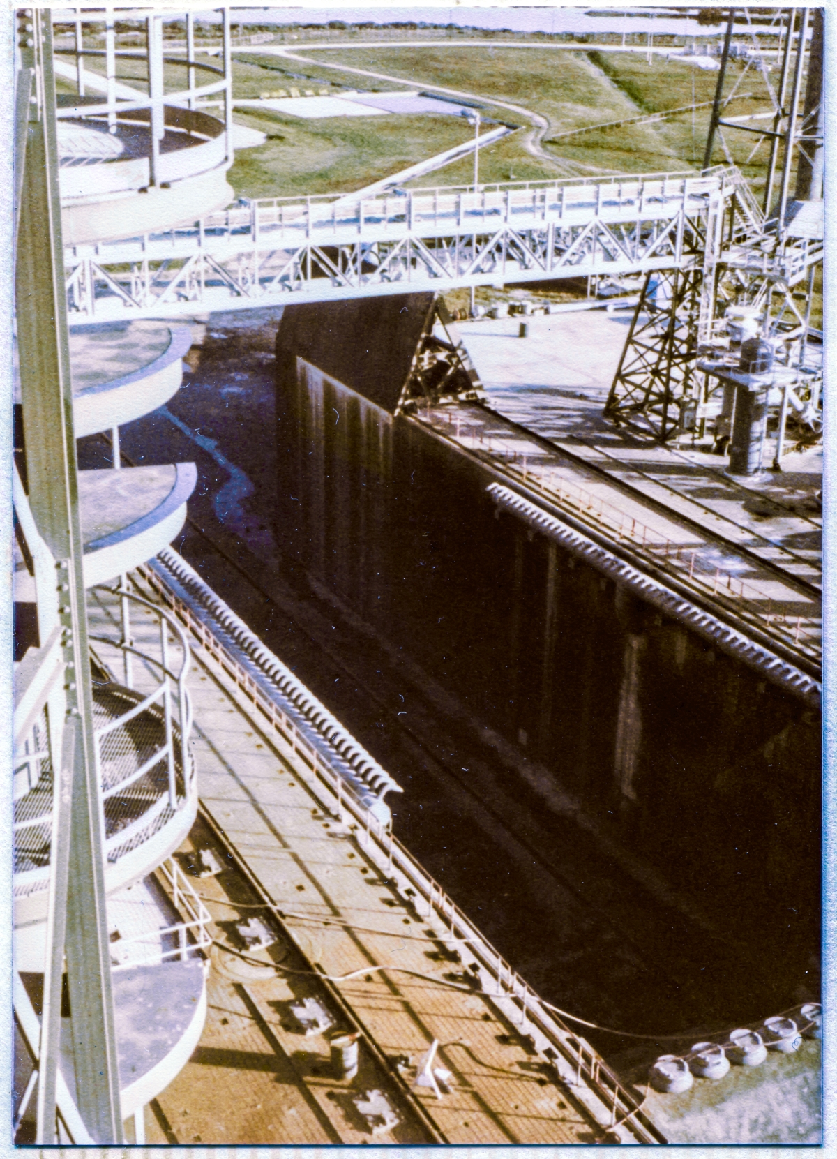 Image 024. From a viewpoint at the front corner of the left Tail Service Mast Access Platform, at the 125’ elevation of the Rotating Service Structure, looking northeast and down, the dark open gash of the Flame Trench at Launch Complex 39-B, Kennedy Space Center, Florida, dominates a frame encompassing a science-fiction array of Ground Support Equipment on and around the raised body of the Pad, surrounding the Flame Trench. Photo by James MacLaren.